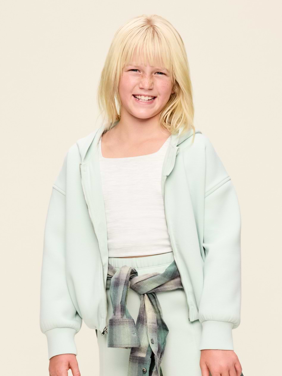 Smiling child in a mint hoodie and matching pants with a white shirt and a plaid shirt tied at the waist, standing against a plain background.