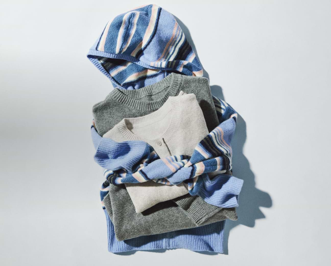 Stack of folded knit sweaters—gray and cream—with a blue striped hoodie draped on top.