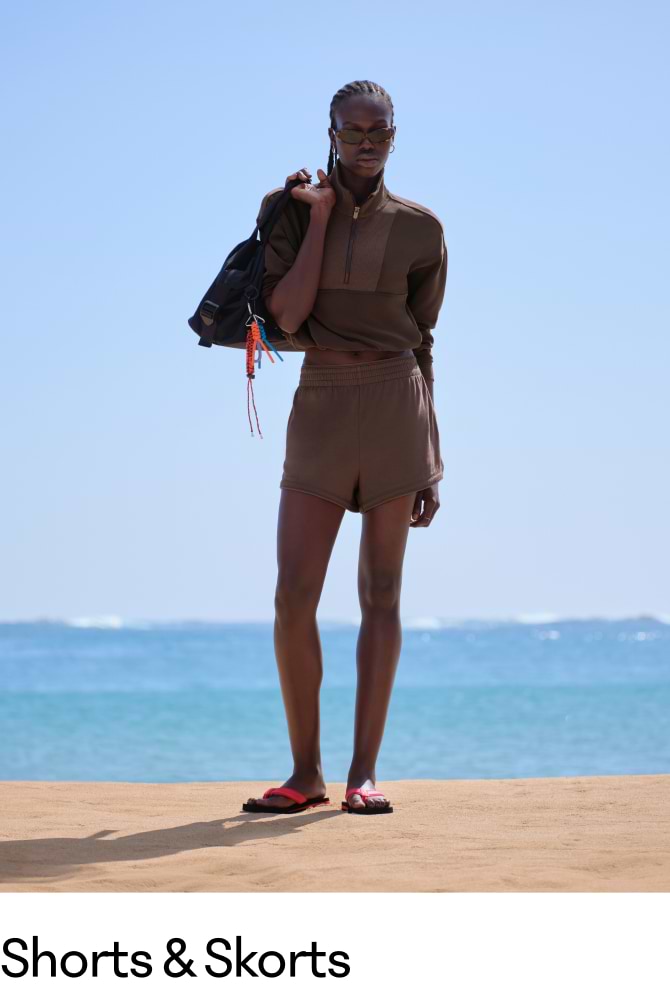 Person in a brown quarter-zip top and matching shorts, wearing sandals and holding a backpack on a beach; text reads “Shorts & Skorts.”