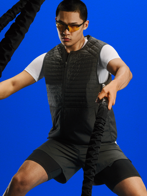 Person in a red athletic T‑shirt over white long sleeves, maroon shorts, white socks and sneakers, wearing mirrored sports sunglasses, standing against a plain beige backdrop.