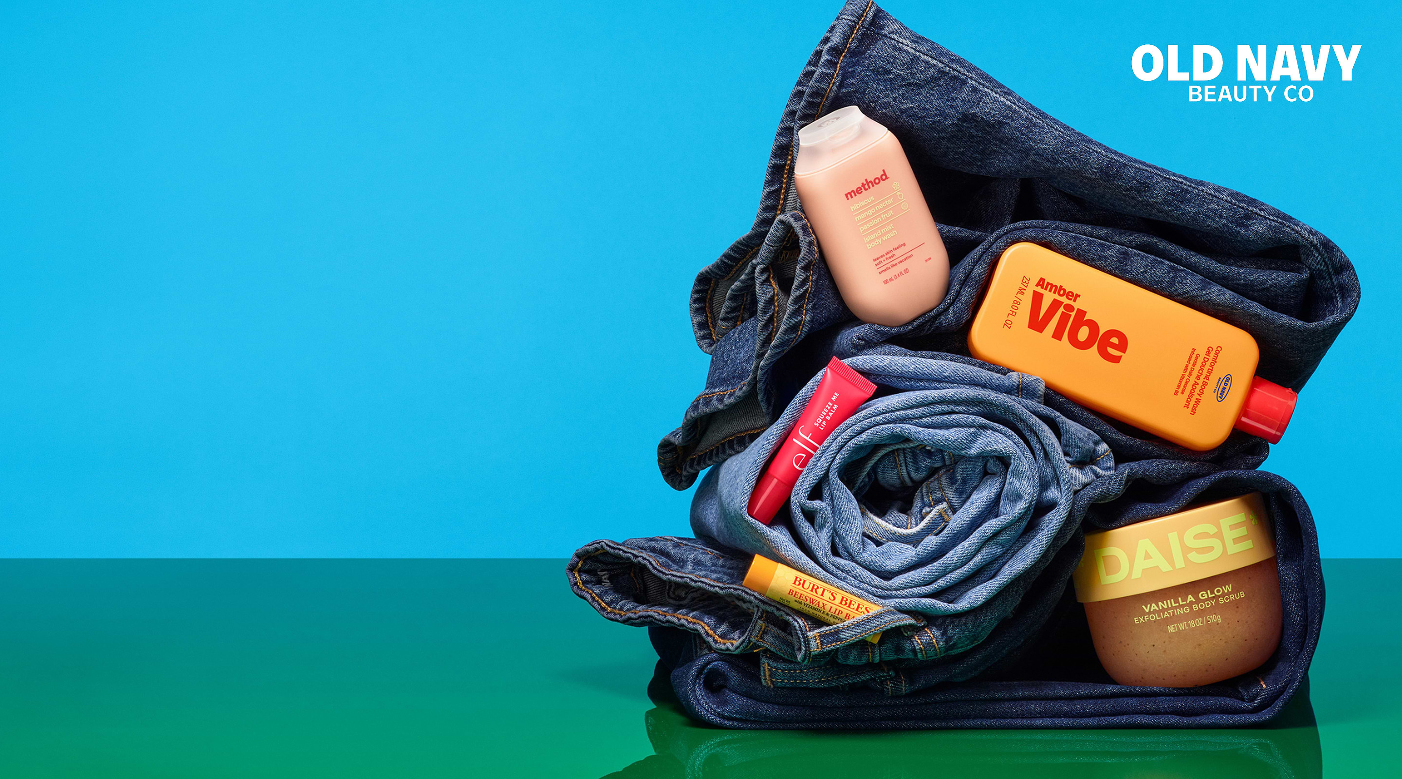 Rolled blue jeans stacked on a green surface with beauty items tucked in—orange Amber Vibe bottle, pink Method wash, e.l.f. gloss, Burt’s Bees balm, and DAISE scrub—under “OLD NAVY BEAUTY CO” on a blue background.