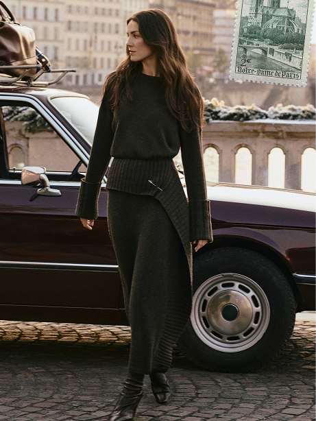 Woman in a dark knit sweater and wrap skirt walks past a maroon classic car on a cobblestone riverside, with a stone balustrade and city buildings in the background.