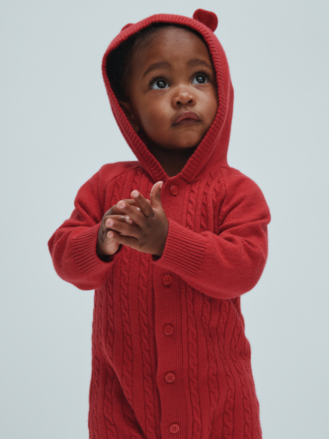 Toddler in a red cable-knit hooded onesie with small bear ears, looking up and clasping hands.
