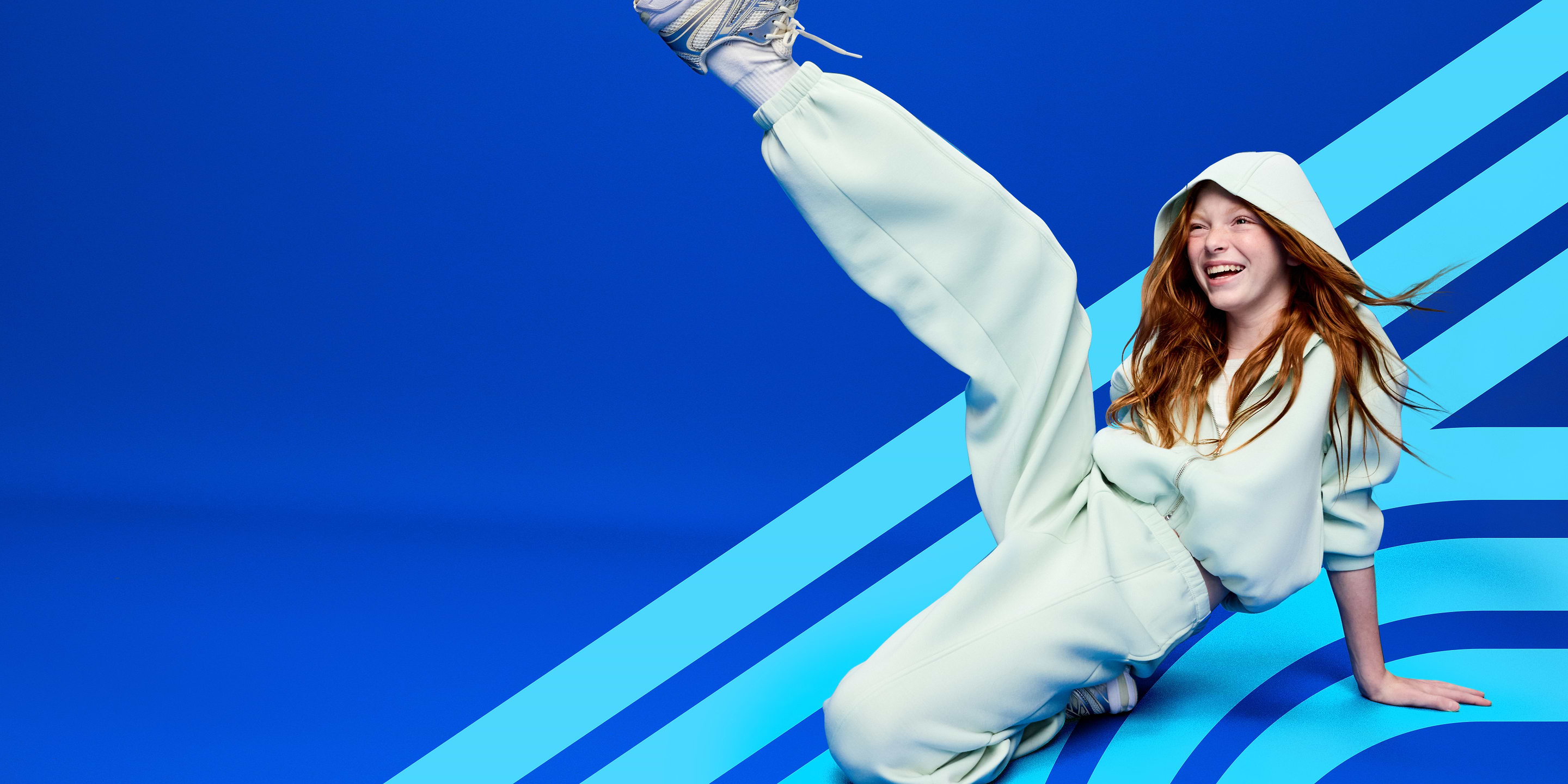 Smiling person in a light mint hoodie and sweatpants kicks one leg up against a blue background with diagonal light-blue stripes.