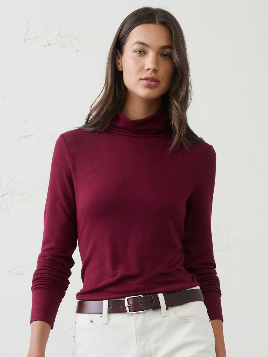 Person wearing a burgundy long-sleeve turtleneck with white jeans and a brown belt, standing against a plain wall.