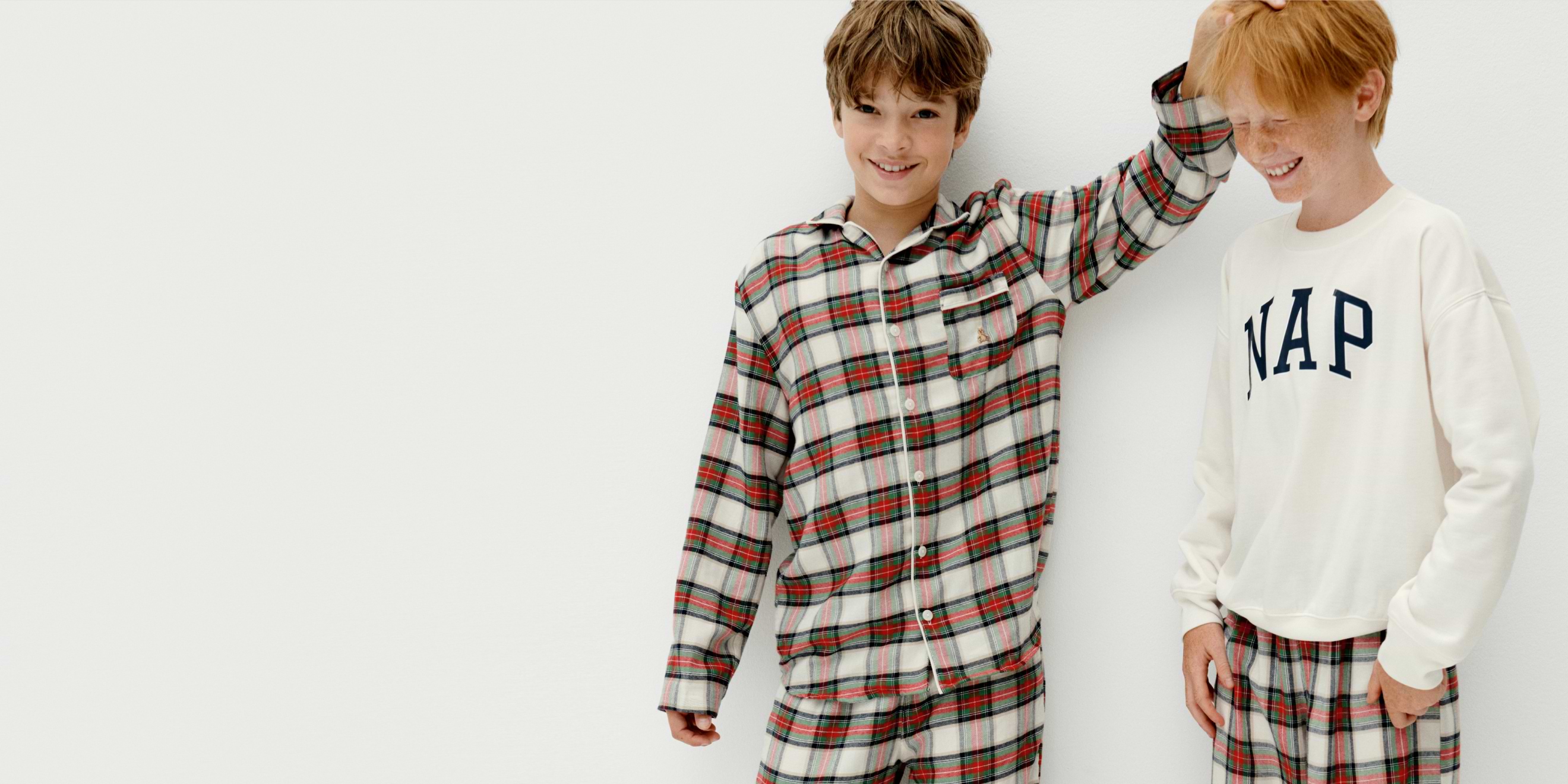 Two smiling kids in plaid pajamas; one ruffles the other’s hair, and one wears a white sweatshirt reading “NAP,” on a white background.