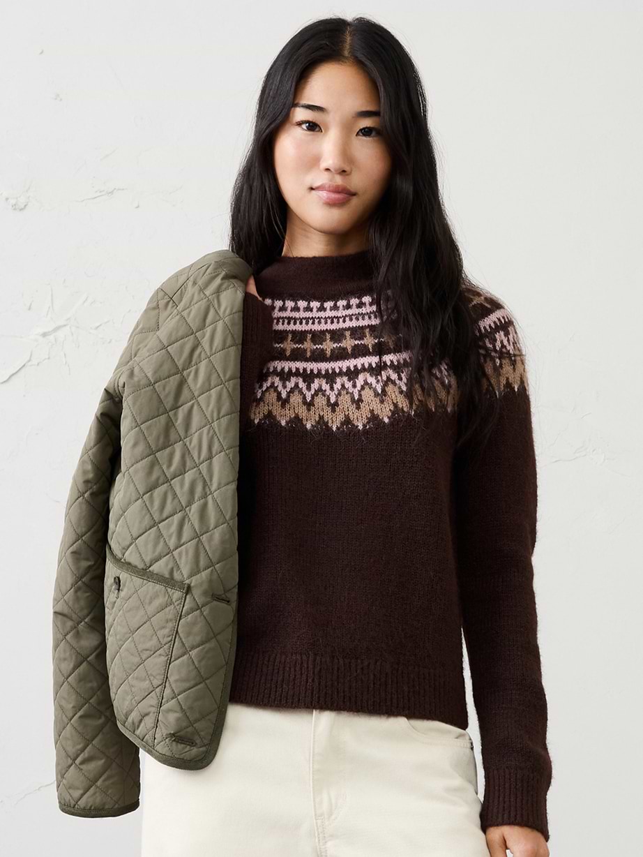 Person in a dark brown Fair Isle sweater with pink/tan yoke, cream pants, and an olive quilted jacket over one shoulder.