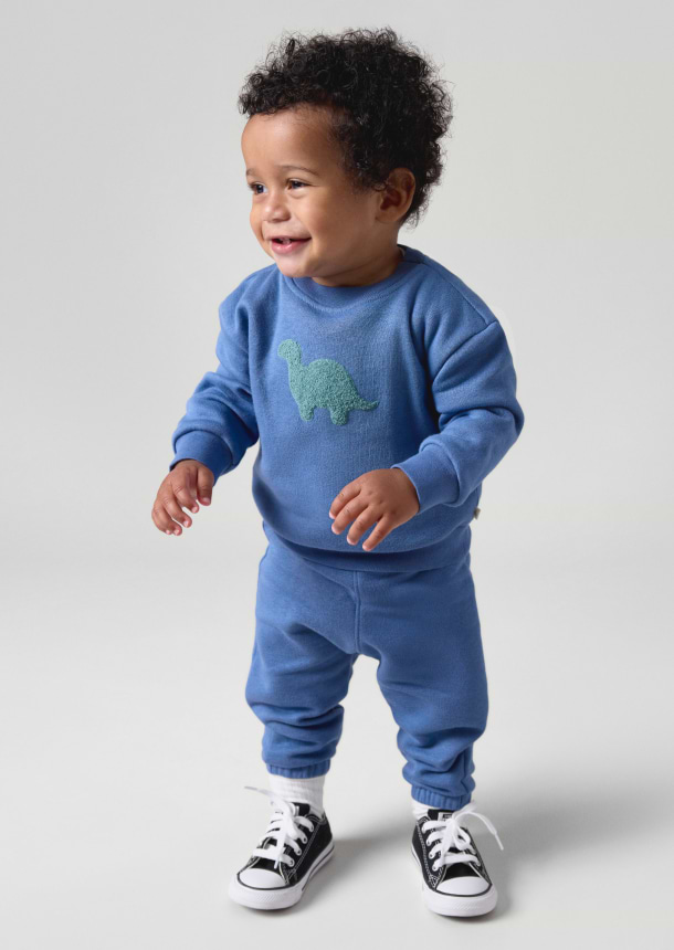 Smiling toddler in a blue dinosaur sweatsuit and black-and-white sneakers, standing.