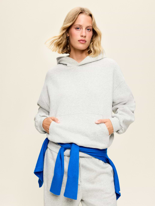 Person in a light gray hoodie and sweatpants with a bright blue sweater tied around their waist, hands in the hoodie pocket, standing against a plain background.