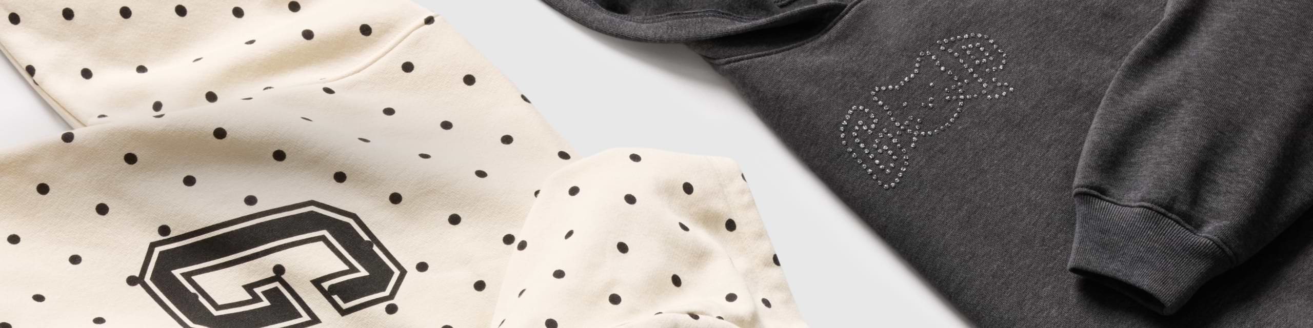 Close-up of two sweatshirts: one cream with black polka dots and a large outlined letter, and one dark gray hoodie with a rhinestone-style bear graphic.