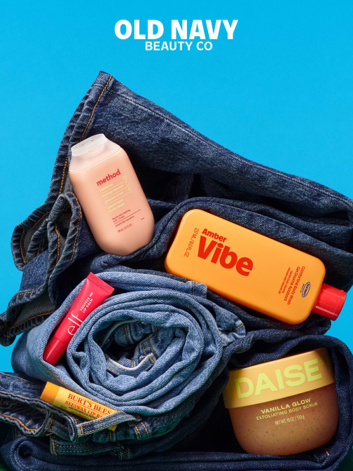 Flat lay of rolled blue jeans on a bright blue background with beauty products—Method body wash, Amber Vibe bottle, e.l.f. lip color, Burt’s Bees balm, and DAISE Vanilla Glow scrub—under “Old Navy Beauty Co” text.