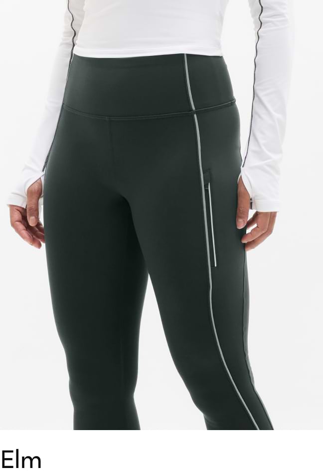 Close-up of a person wearing dark green high-waisted athletic leggings with reflective piping and a side zip pocket, paired with a white long-sleeve top.