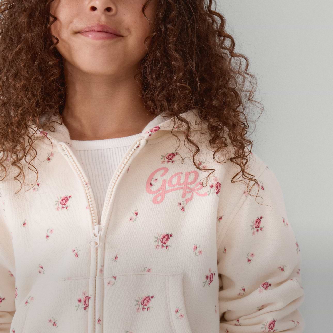 Close-up of a child with curly hair wearing a cream floral zip-up hoodie with a pink “Gap” logo.