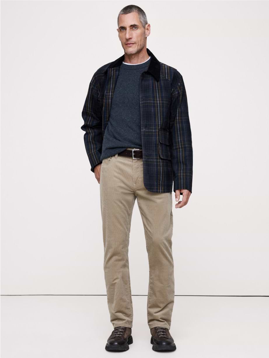 Person wearing a dark plaid jacket over a gray sweater with tan corduroy pants and brown shoes, standing against a plain white background.