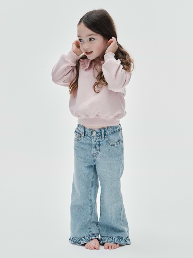 Child in a pink sweatshirt and light blue flared jeans stands barefoot, hands near ears, looking to the side.