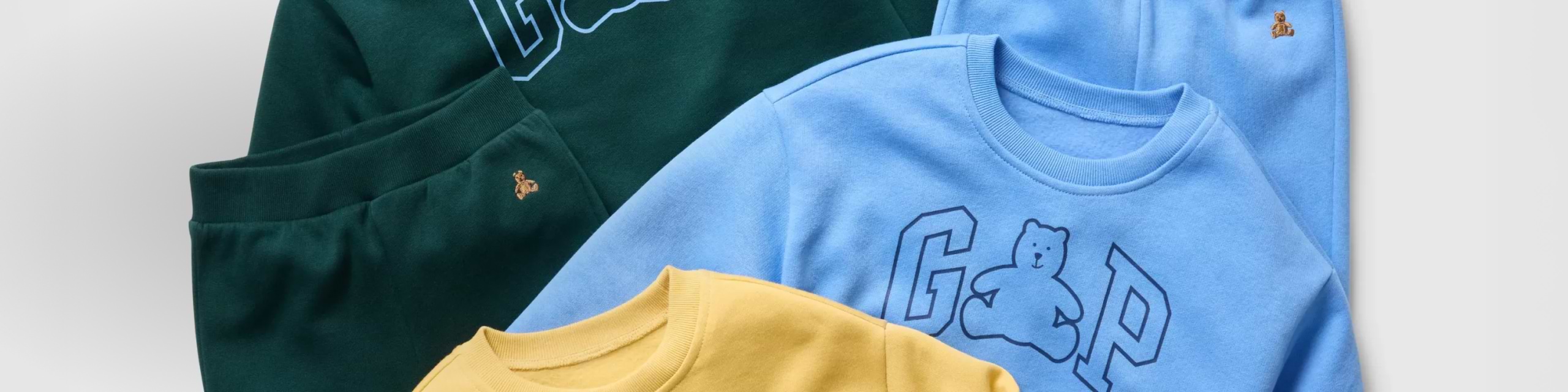 Folded GAP sweatshirts and joggers in green, light blue, and yellow, featuring teddy bear logos.