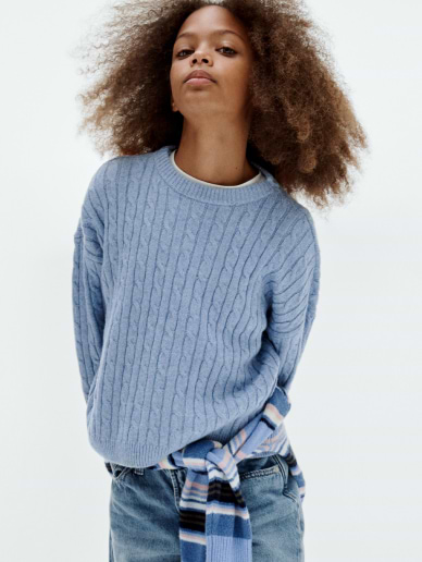 Young person standing with hands behind their back, wearing a light blue cable-knit sweater and jeans, with a striped scarf tied at the waist against a plain background.