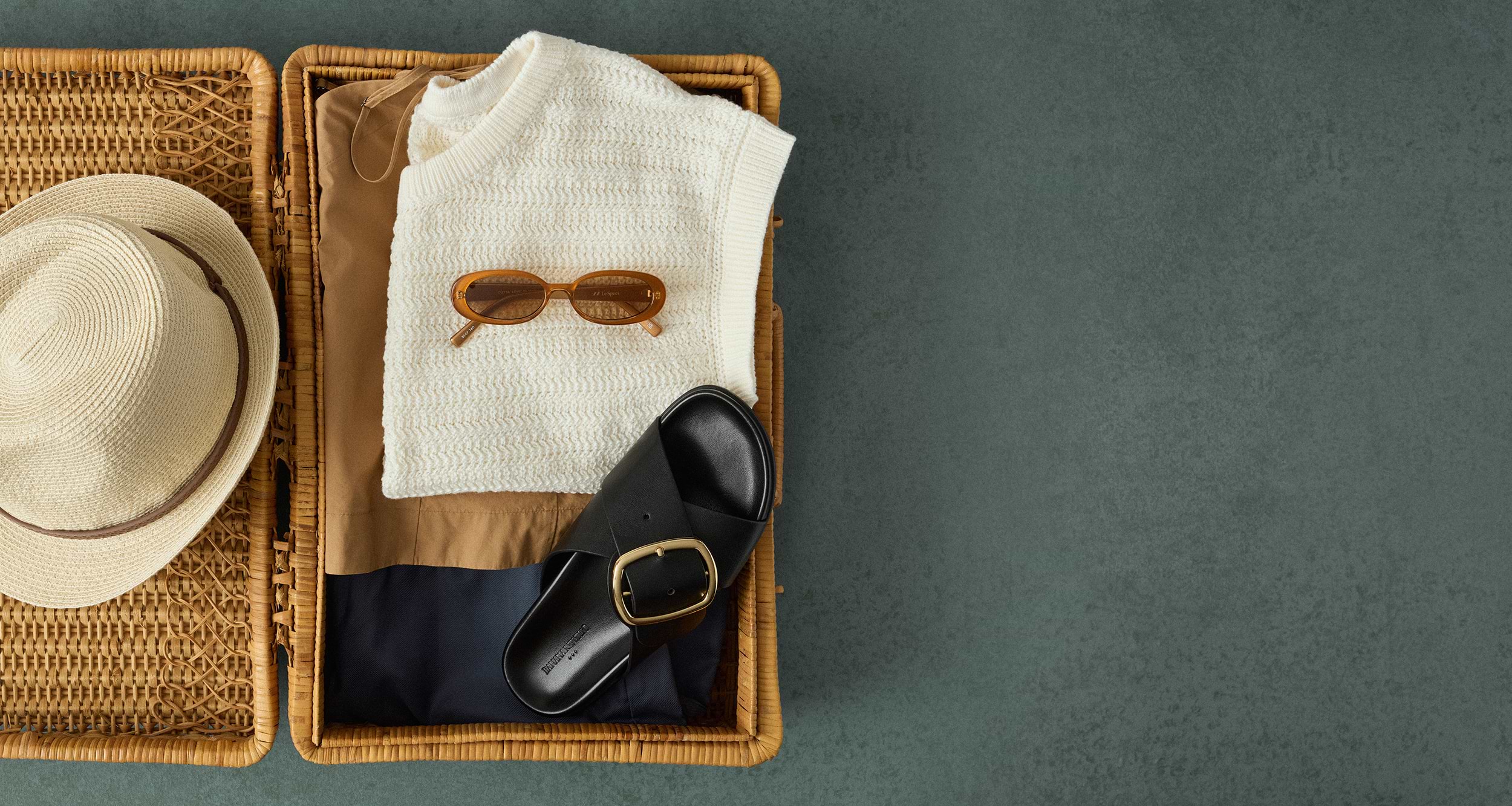 Open wicker suitcase with a straw hat, cream knit top, brown pants, sunglasses, and a black sandal.