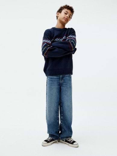 Person standing with arms crossed, wearing a navy sweater with colorful patterned sleeves, wide-leg blue jeans, and black sneakers against a plain white background.