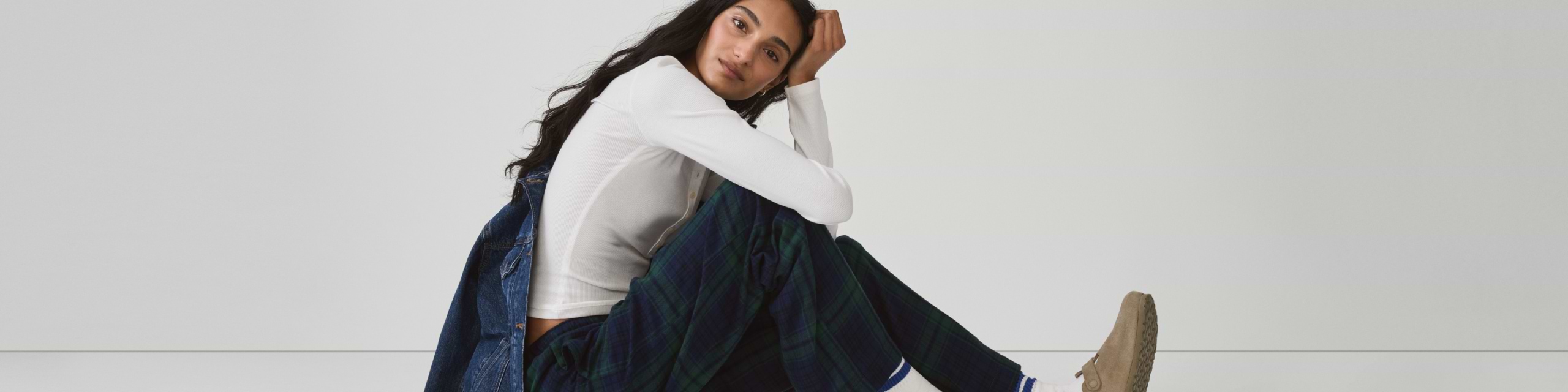 Person seated on the floor, resting head on hand, wearing a white long-sleeve top, dark plaid pants, and tan shoes against a plain background.