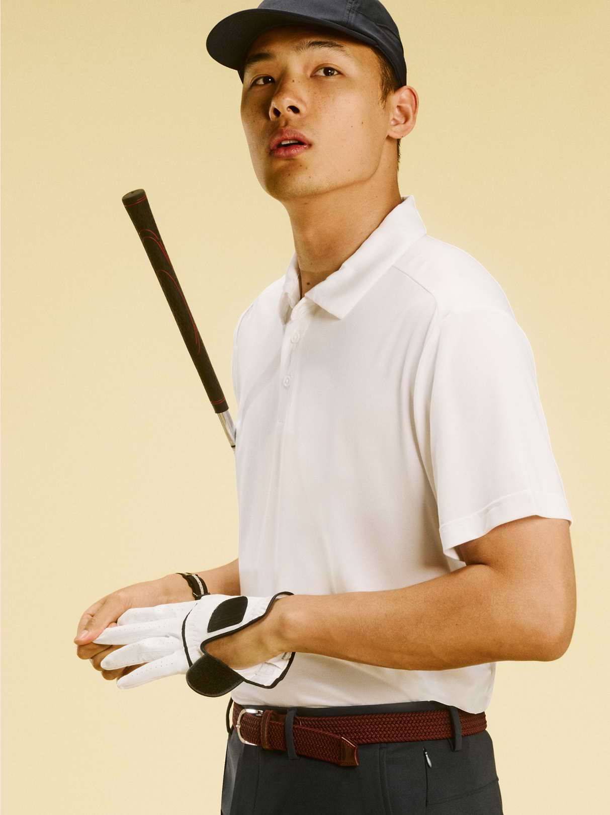 Person in a white polo and cap putting on a golf glove, with a club resting on their shoulder against a light background.