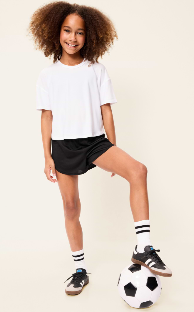 Smiling child in a white T-shirt and black shorts standing with one foot on a soccer ball.