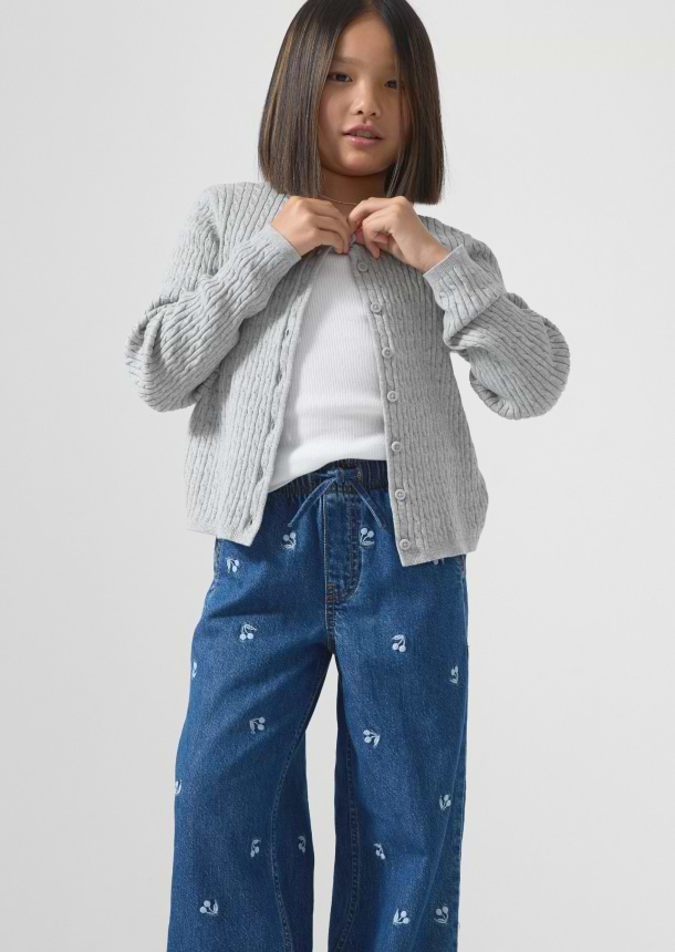 Child buttoning a gray cable‑knit cardigan over a white top, wearing wide‑leg blue jeans with small floral prints.