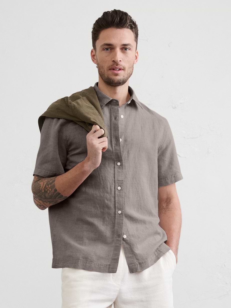 Model wearing a gray short‑sleeve button-up linen shirt and white pants, jacket over shoulder.