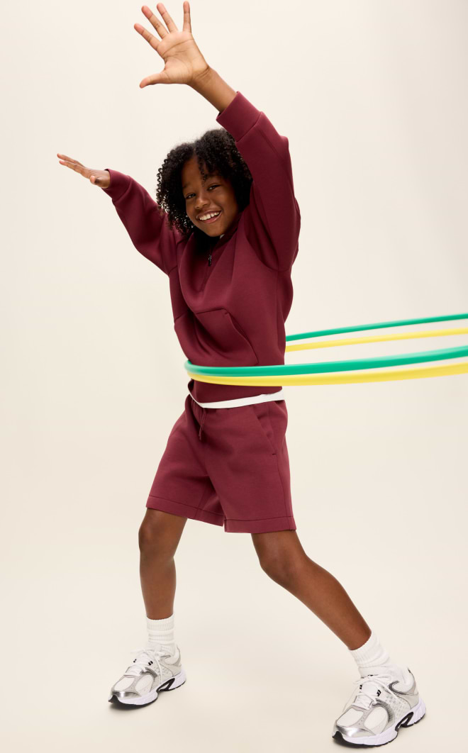 Smiling child in a maroon sweatshirt and shorts spins green and yellow hula hoops around their waist, arms raised, on a plain background.