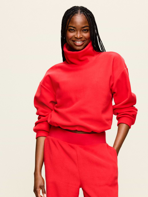 Smiling person with long braids wearing a bright red turtleneck sweatshirt and matching pants, hand in pocket against a plain background.