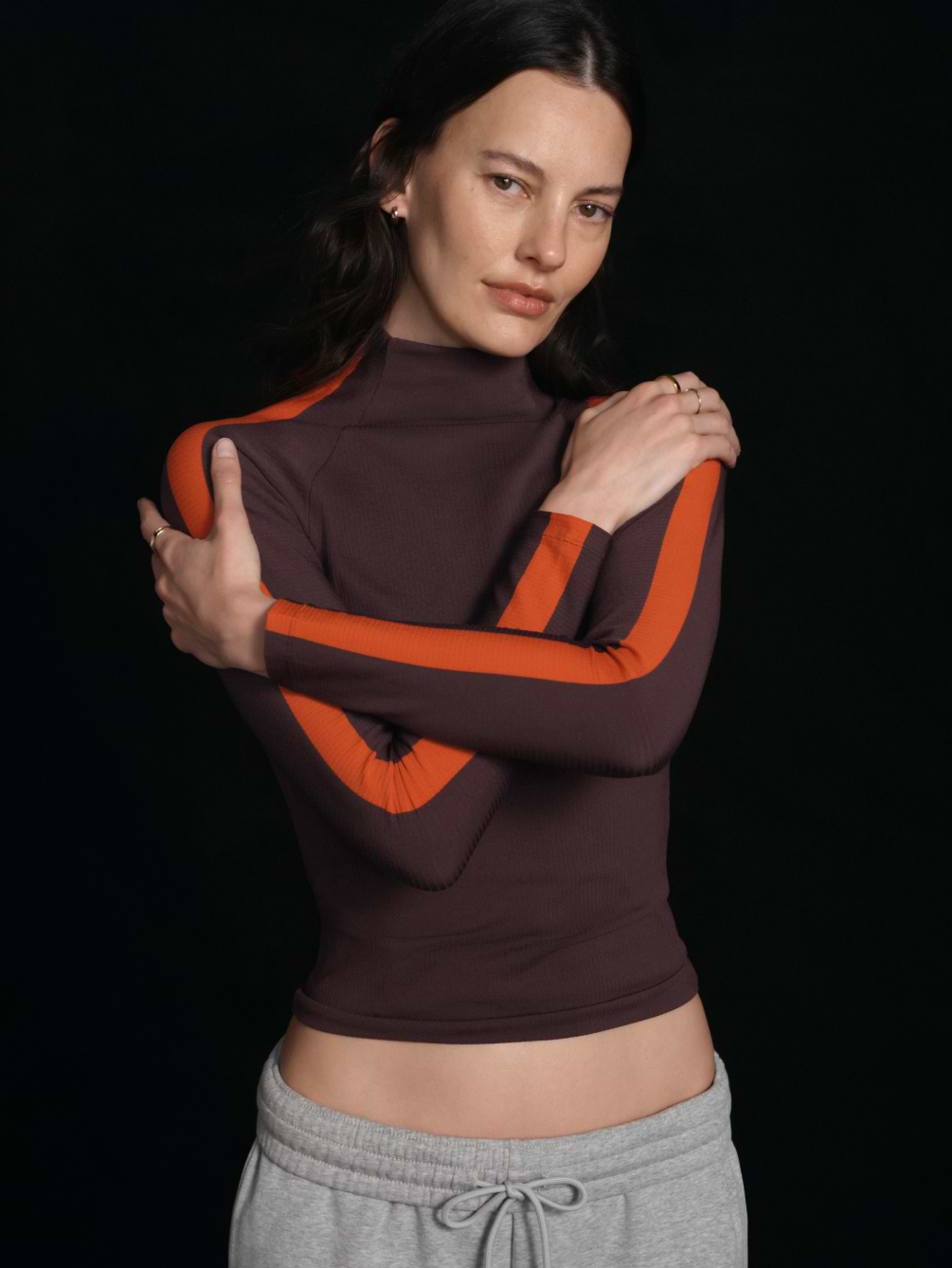 Person in a fitted brown top with orange striped sleeves, arms crossed, on a black background.