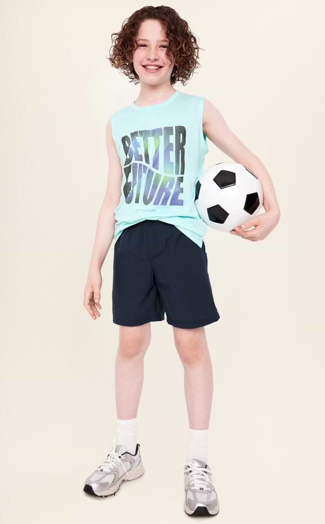 Smiling child with curly hair holds a soccer ball, wearing a mint sleeveless “BETTER FUTURE” shirt, navy shorts, and sneakers against a plain background.