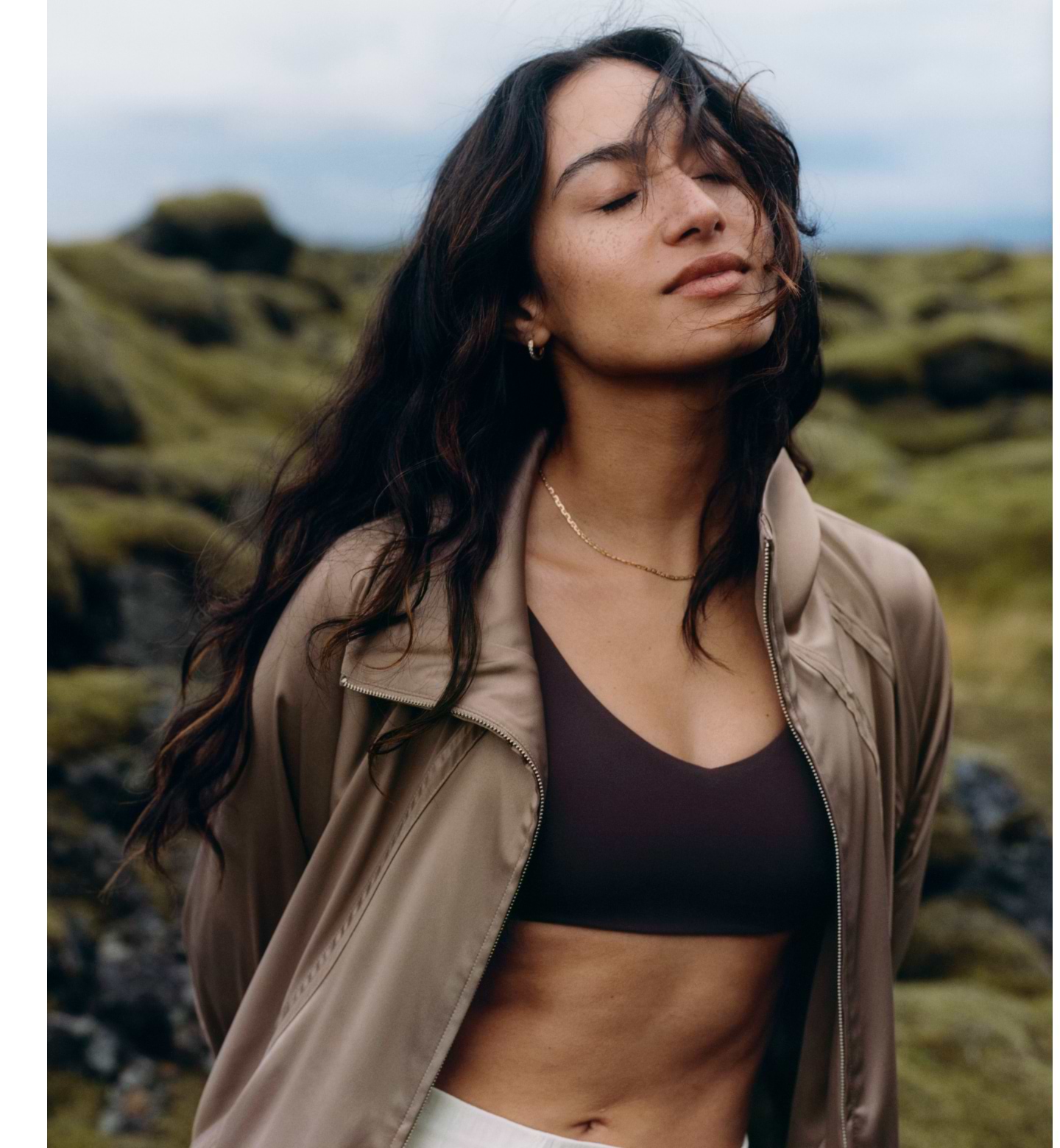 Person with long wavy hair, eyes closed, wearing a sports bra and light jacket, standing among mossy rocks with wind blowing.