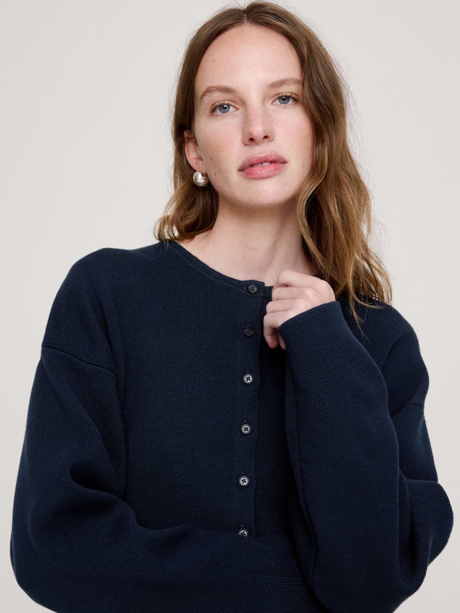 Person wearing a dark navy button-front cardigan, lightly touching the neckline, facing the camera against a plain light background.