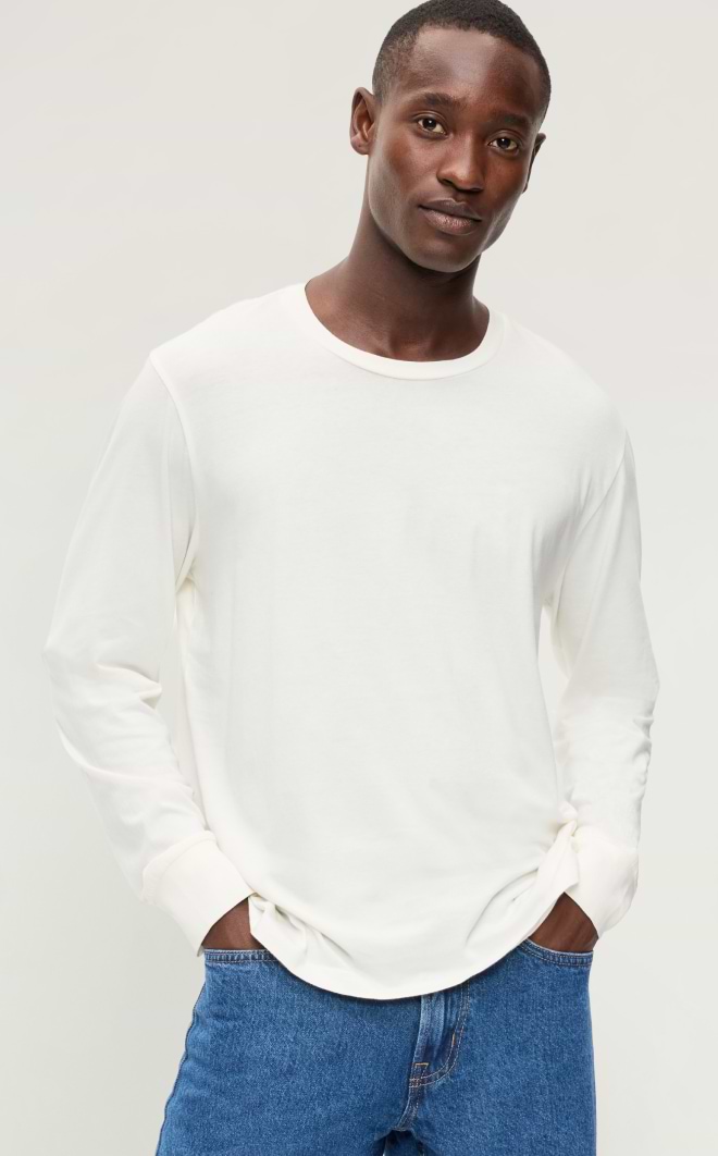 Person wearing a white long-sleeve shirt and blue jeans, hands in pockets, standing against a plain background.